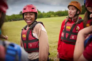 A group of children wearing helmets and life jackets stand together outdoors on a grassy field, with one child smiling at the camera.