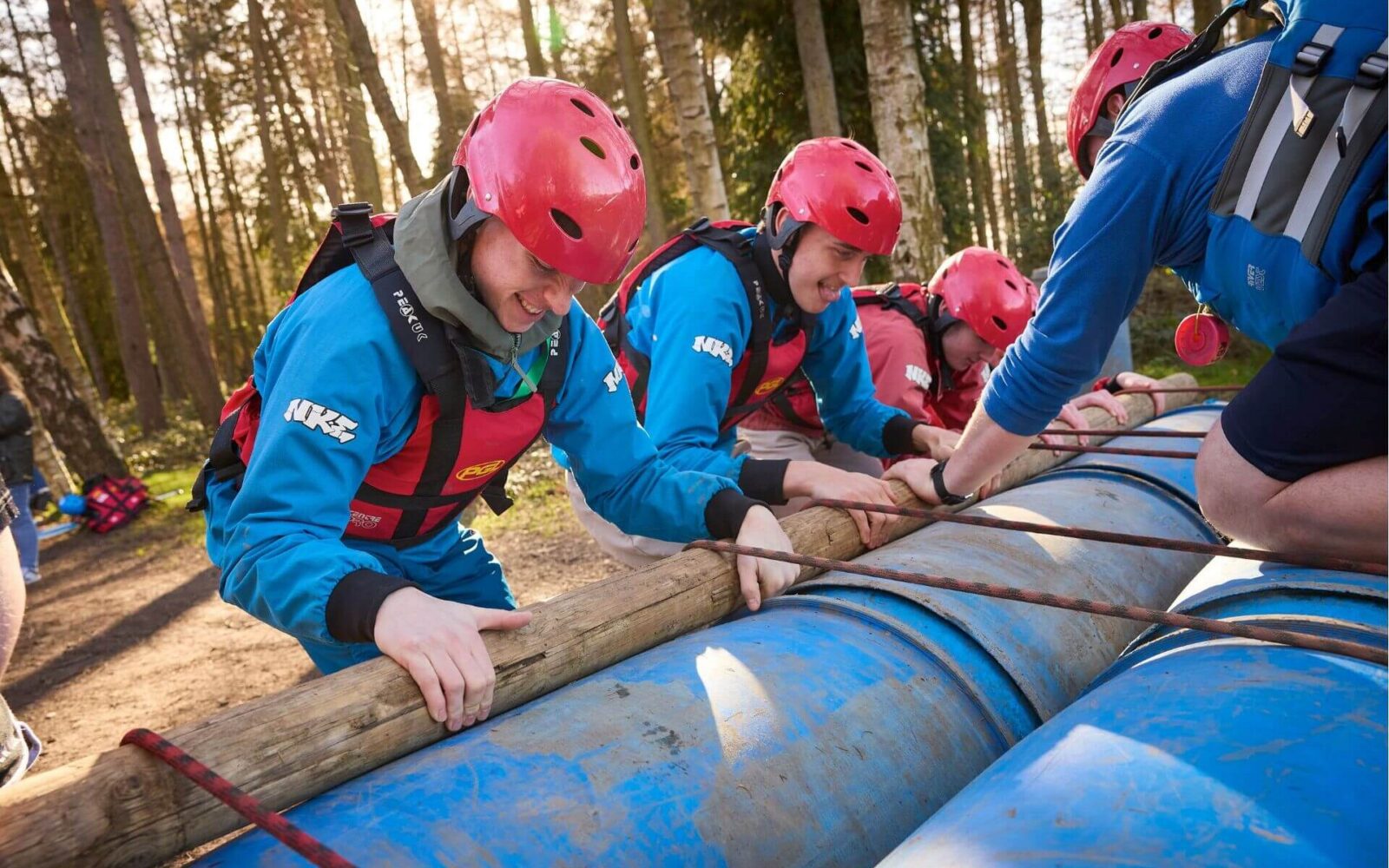 Young people wearing helmets and buoyancy aids working together to build a raft using logs and barrels during a PGL SEND residential trip