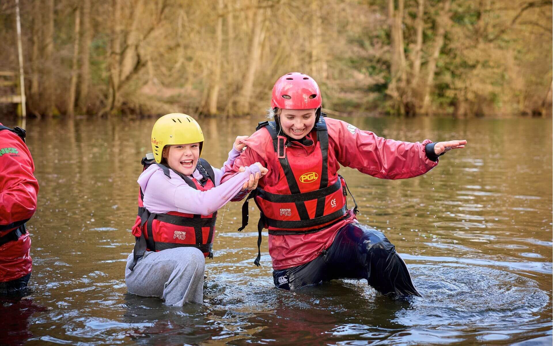 Two laughing young people wearing helmets and buoyancy aids wading through a river together during a PGL SEND residential trip