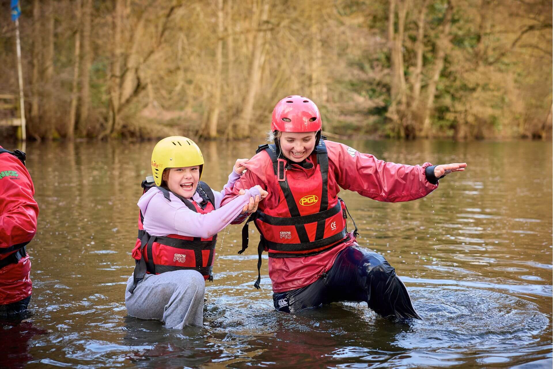 Two laughing young people wearing helmets and buoyancy aids wading through a river together during a PGL SEND residential trip