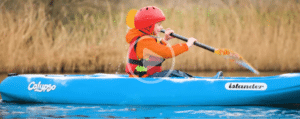 Child wearing a red helmet and orange jacket paddles a blue kayak labelled "Calypso Islander" on calm water with tall grass in the background.