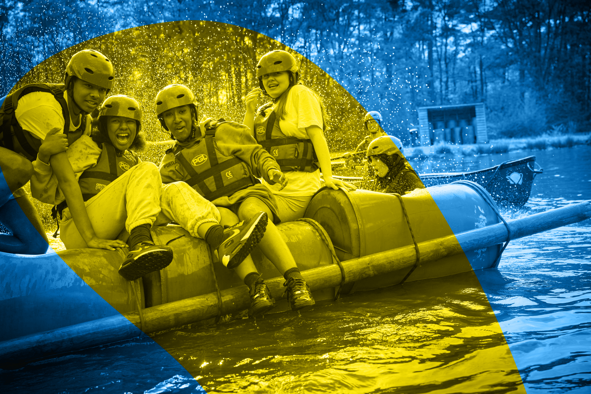 Four people in helmets and life jackets sit on a makeshift raft in a lake, smiling and splashing water, with trees and a small building visible in the background.