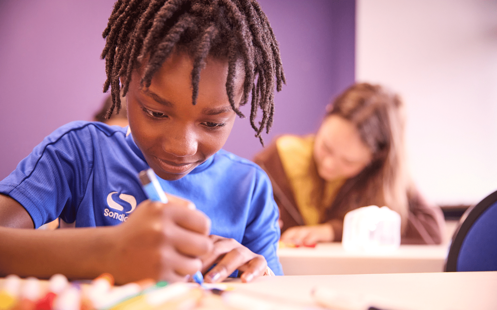 Child in blue shirt drawing with felt-tip pens at a desk, with another pupil working in the background in a classroom setting.