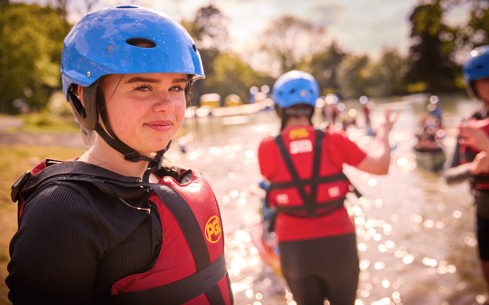 Young person in a blue helmet and red life vest stands near water, with other people in similar kit in the background. It appears to be a group outdoor water activity.