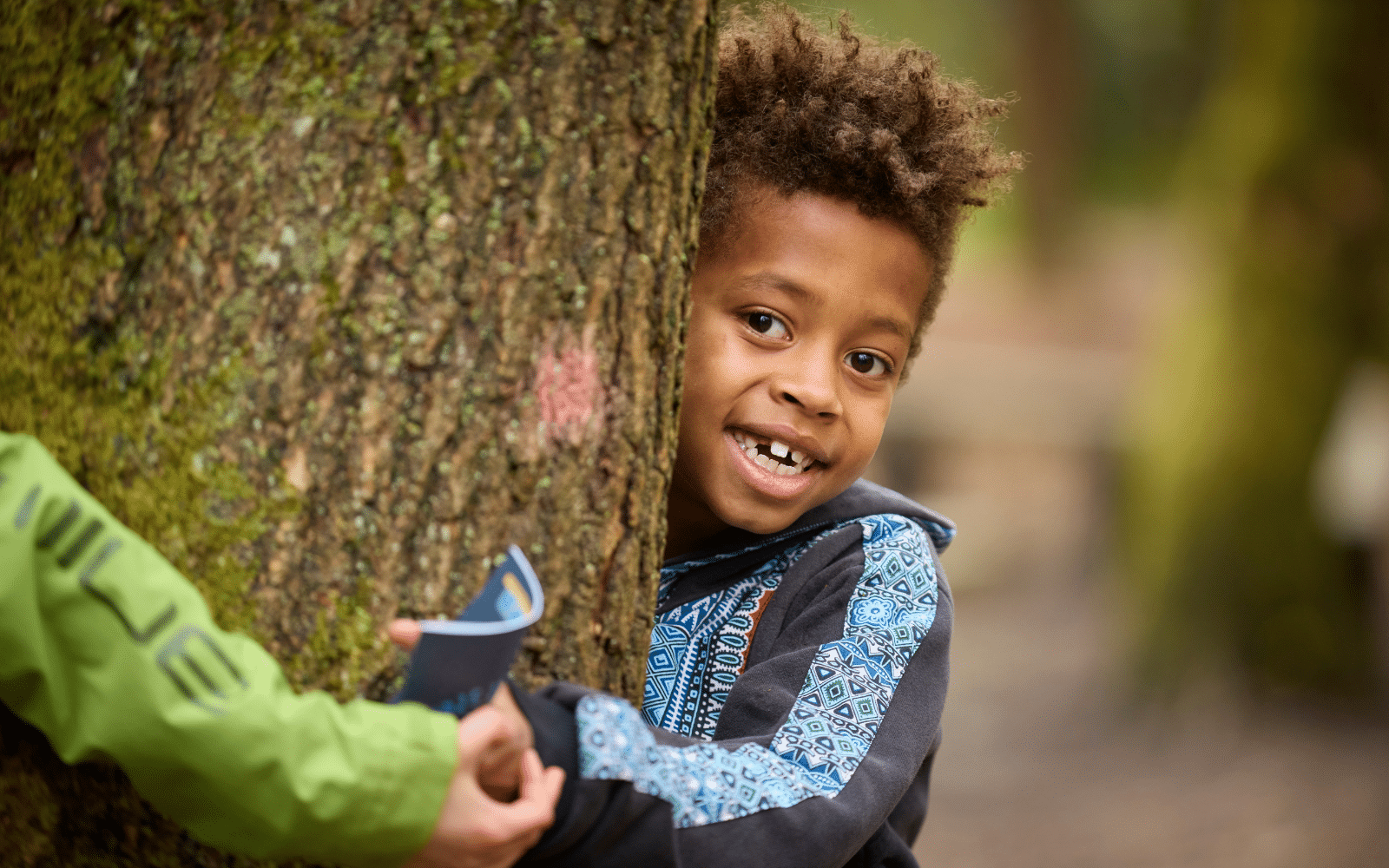 A child with curly hair hugs a tree and smiles. Someone in a green jacket extends an object towards the child. The background is a forest setting.