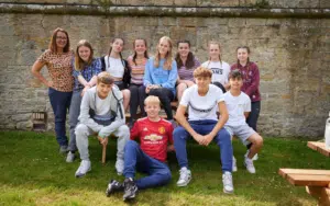 A group of teenagers and an adult sit and stand together on grass in front of a stone wall, posing for a group photo outdoors during one of their Curriculum for Excellence school trips.
