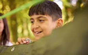A boy smiles whilst holding a green tarpaulin outdoors, with blurred trees in the background—capturing the fun and hands-on learning often experienced during Curriculum for Excellence school trips.