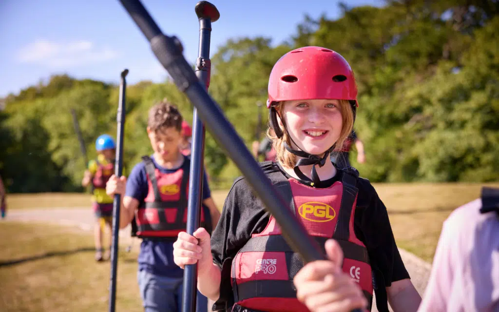 Children wearing helmets and life jackets hold paddles outdoors on a sunny day, preparing for an activity as part of inspiring Curriculum for Excellence school trips.