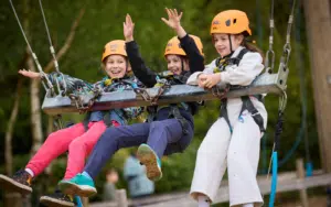 Three children in helmets and harnesses sit on a metal swing outdoors, smiling and raising their arms, with trees and a wooden fence in the background—a perfect moment capturing the spirit of Curriculum for Excellence school trips.