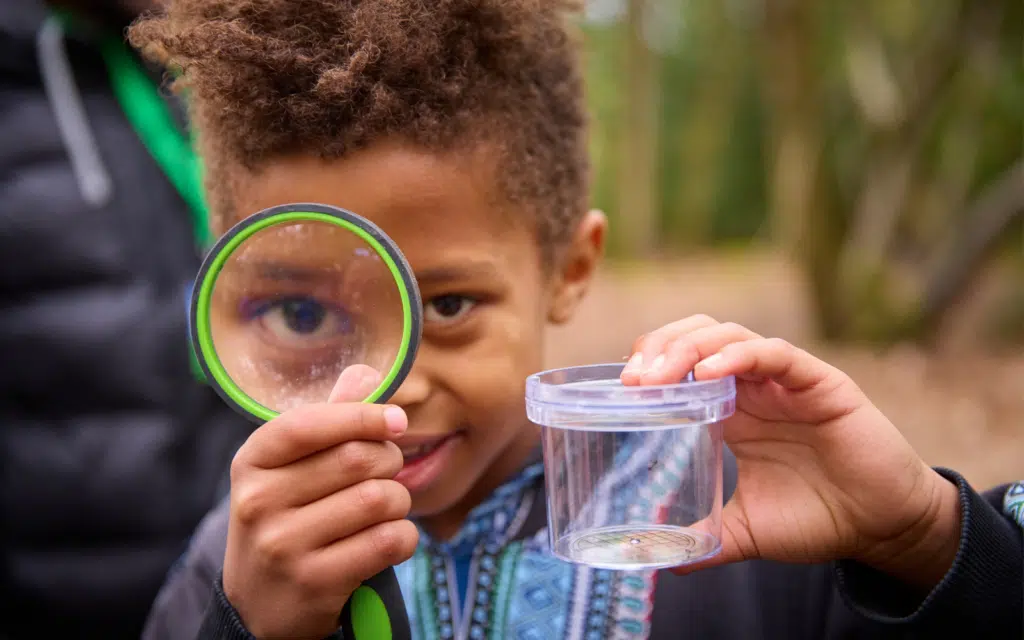 A child holds a magnifying glass to one eye and an empty clear container in the other hand, standing outdoors—an engaging moment that captures the spirit of Curriculum for Excellence school trips.