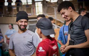 Three young men stand together indoors, smiling and talking. One wears a beanie, one holds a mobile, and a younger boy in a red shirt faces them—perhaps discussing their latest Curriculum for Excellence school trips. Other people are blurred in the background.