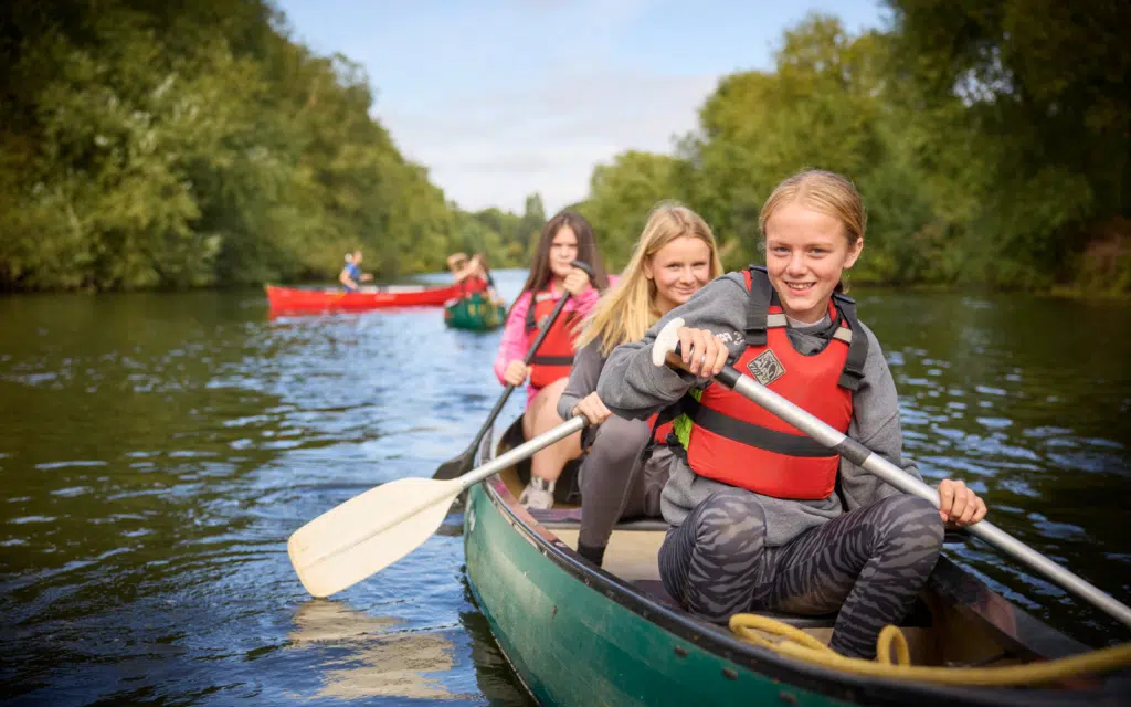 Three girls wearing red life jackets paddle a canoe on a calm river, with other canoes and green trees visible in the background—a perfect example of the adventurous spirit encouraged on Curriculum for Excellence school trips.