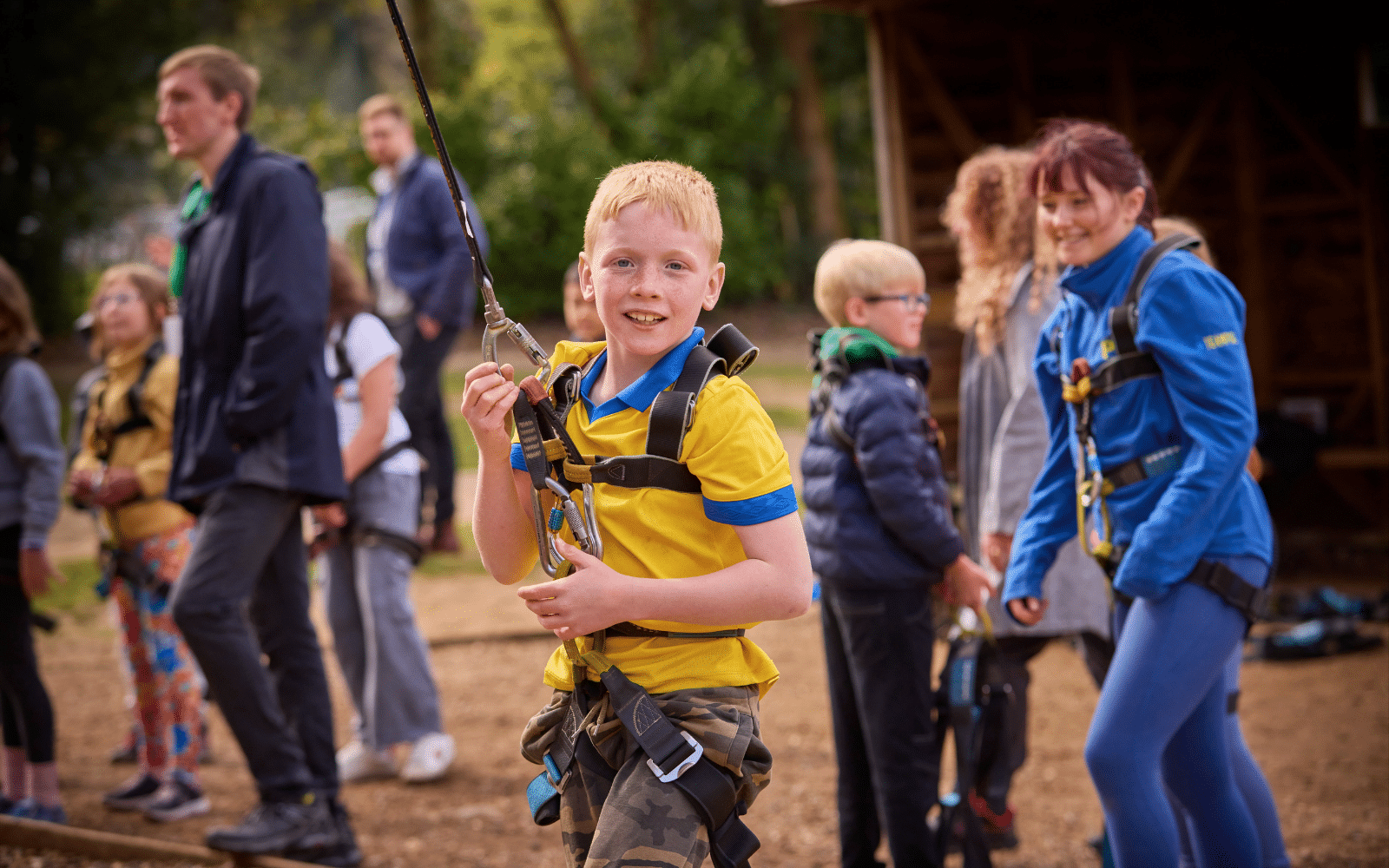A boy in a yellow shirt wearing a harness smiles at the camera outdoors, with other children and adults standing in the background.