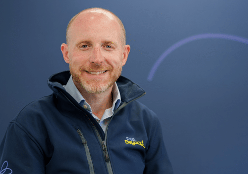 A man with a beard and short hair smiles at the camera, wearing a navy jacket with a “PCI Beyond” logo, in front of a plain blue background.