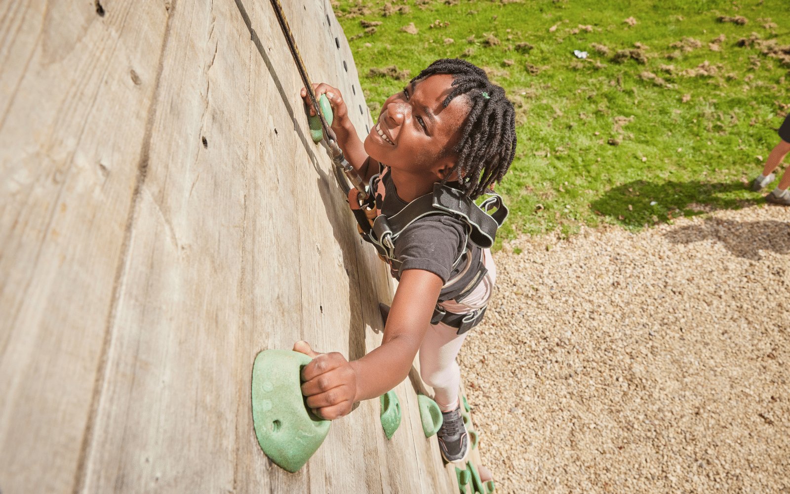 A child wearing a harness climbs a wooden outdoor climbing wall, gripping green handholds and looking upwards.