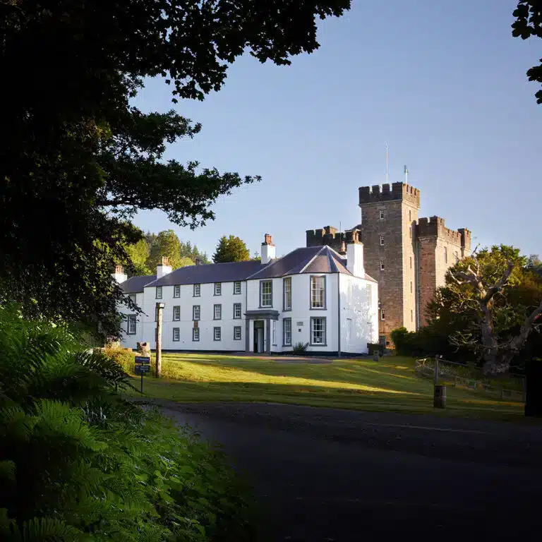 A white manor house stands beside a stone castle tower, surrounded by grass, trees, and a tarmacked road in daylight.