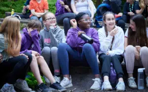 A group of young people sit outdoors on benches, smiling and laughing, with trees in the background. Several wear purple hoodies and casual clothes. One person holds a water bottle.