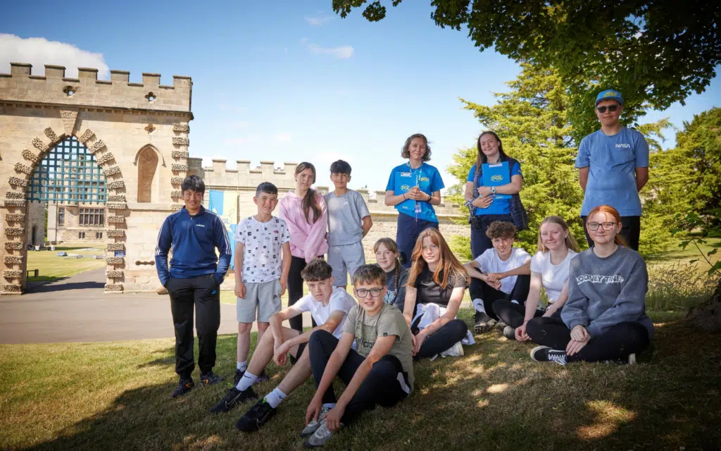 A group of teenagers poses on grass in front of a stone building with an arched gate on a sunny day. Some sit whilst others stand, with trees and blue sky in the background.