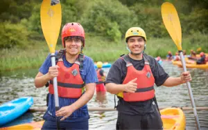 Two people wearing helmets and life jackets stand by kayaks on a river, holding paddles and smiling. Other kayakers are visible in the background. Trees and greenery surround the water.