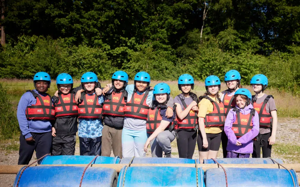 A group of people wearing blue helmets and red life vests stand side by side outdoors behind blue barrels, with trees in the background.