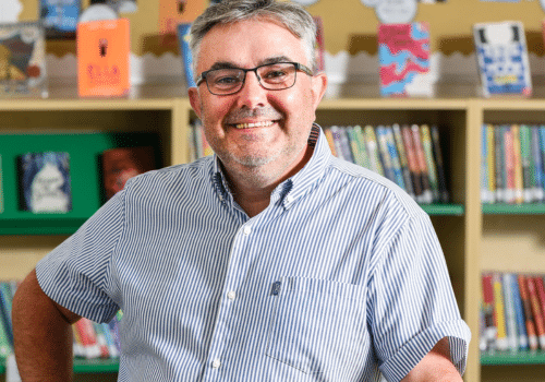 A man with glasses and grey hair, wearing a striped shirt, stands smiling in front of shelves filled with books in a library.