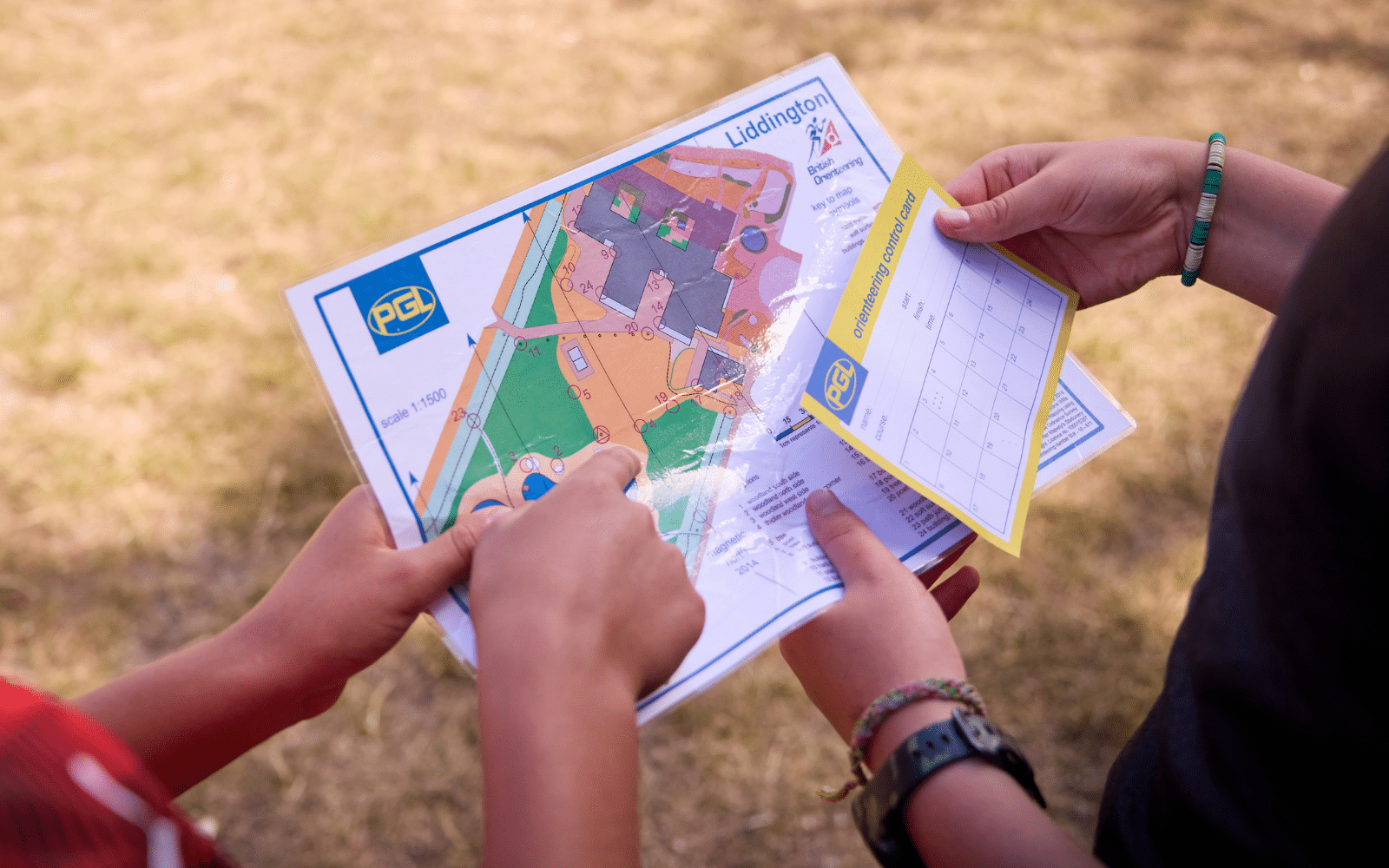 Two people hold an orienteering map and control card at Liddington, with one person pointing to a specific location on the map.