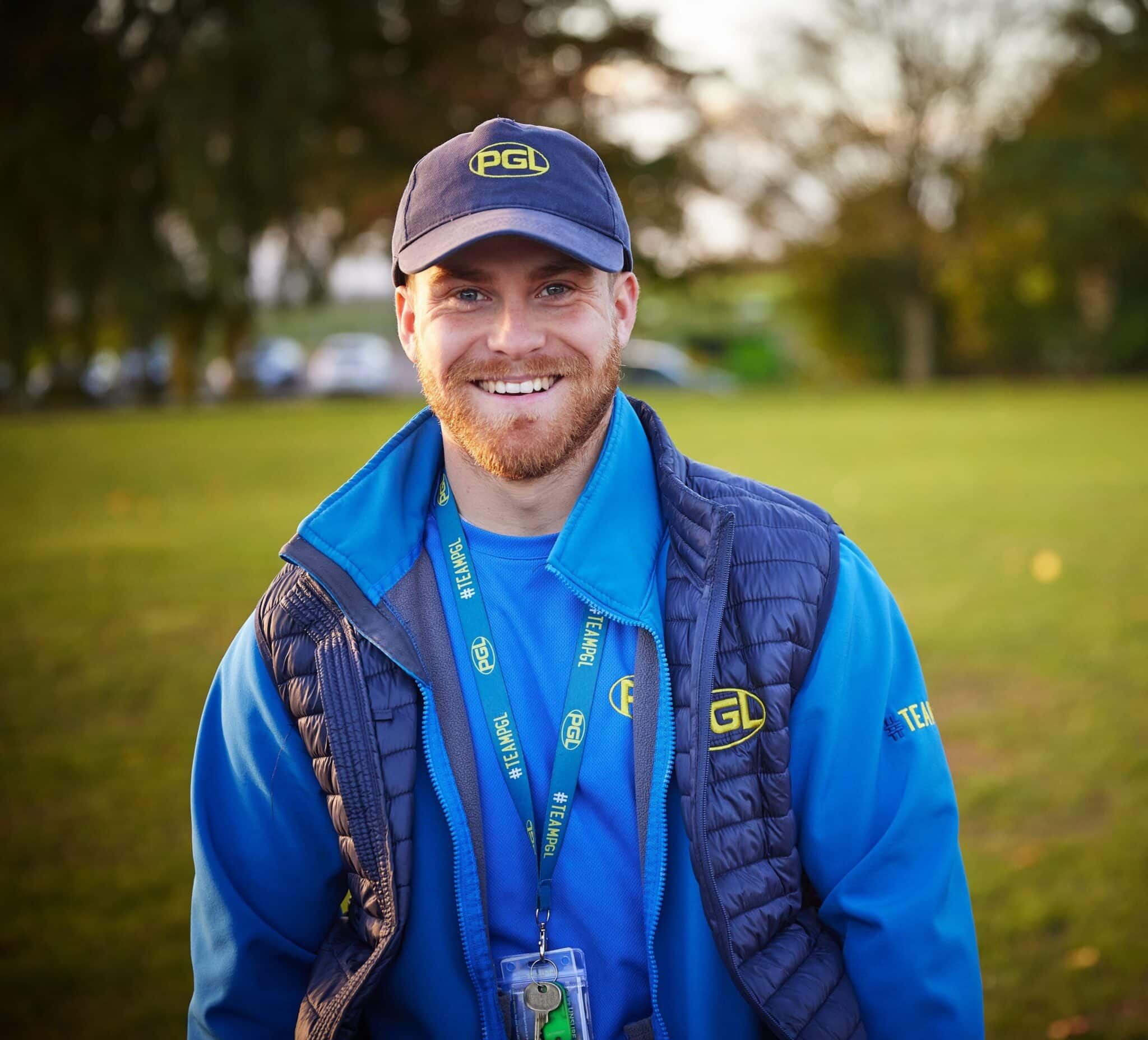 A man wearing a blue PGL uniform and cap stands outside on grass, smiling at the camera with trees and parked cars in the background.