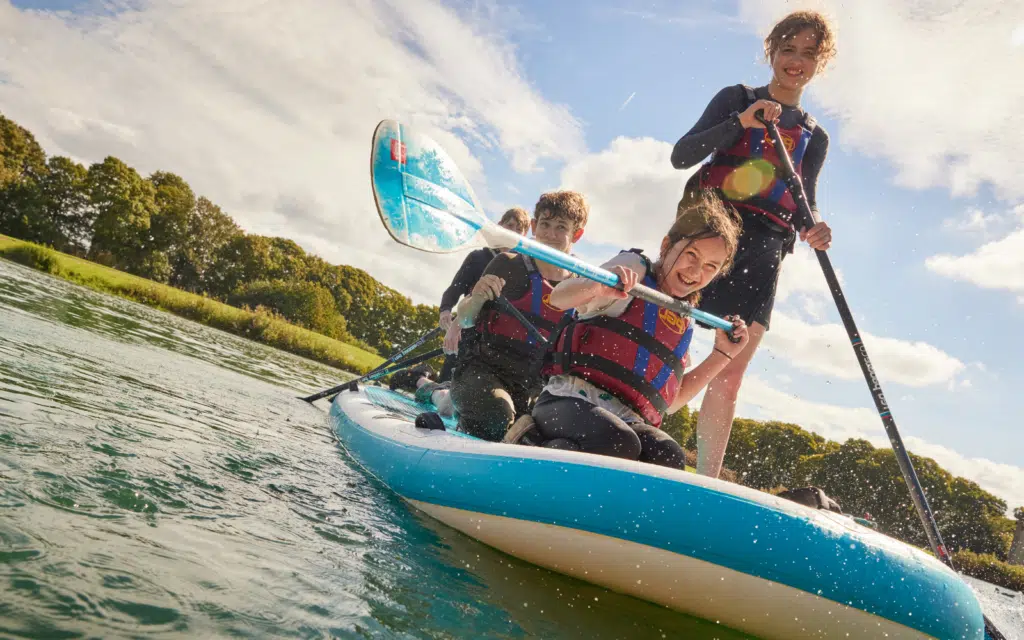 Four people wearing life jackets THRIVE! as they paddle together on a blue inflatable kayak along a river, surrounded by greenery and under a partly cloudy sky.