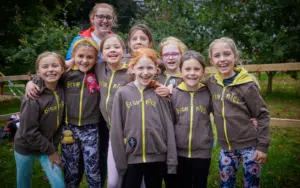 Nine girls in brown “Brownies” hoodies stand together outdoors, smiling at the camera with trees and a wooden fence in the background, ready to THRIVE!.
