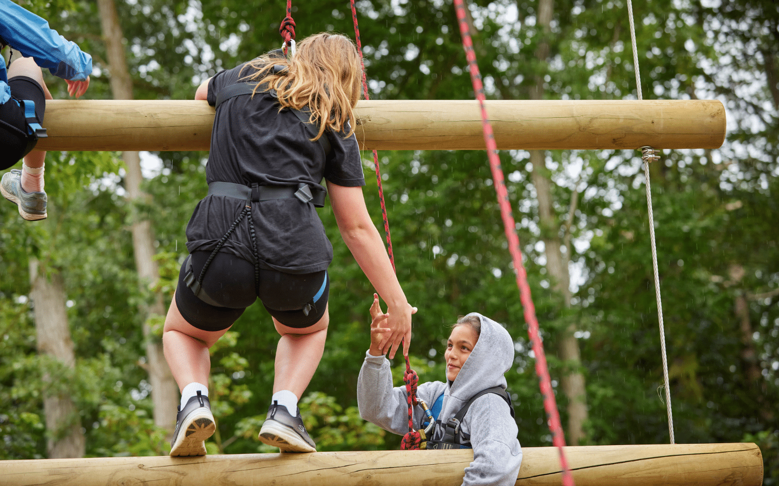 Two people wearing harnesses on a ropes course; one helps the other climb a wooden beam in an outdoor, wooded setting.