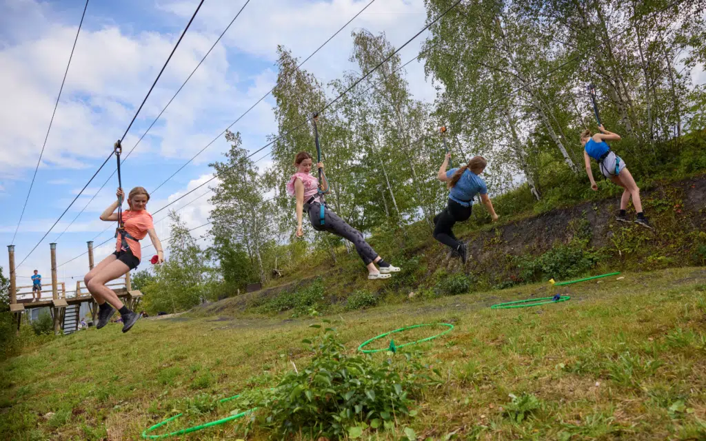Four people wearing harnesses ride zip wires parallel to each other above a grassy slope dotted with green hoops, surrounded by trees and a partly cloudy sky—a thrilling experience that invites adventurers to UNITE! amidst nature’s beauty.
