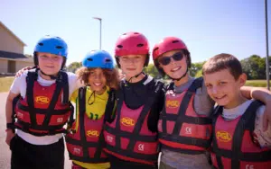 Five children wearing life jackets and helmets stand close together outdoors, smiling at the camera on a sunny day, ready to UNITE! for fun and adventure.