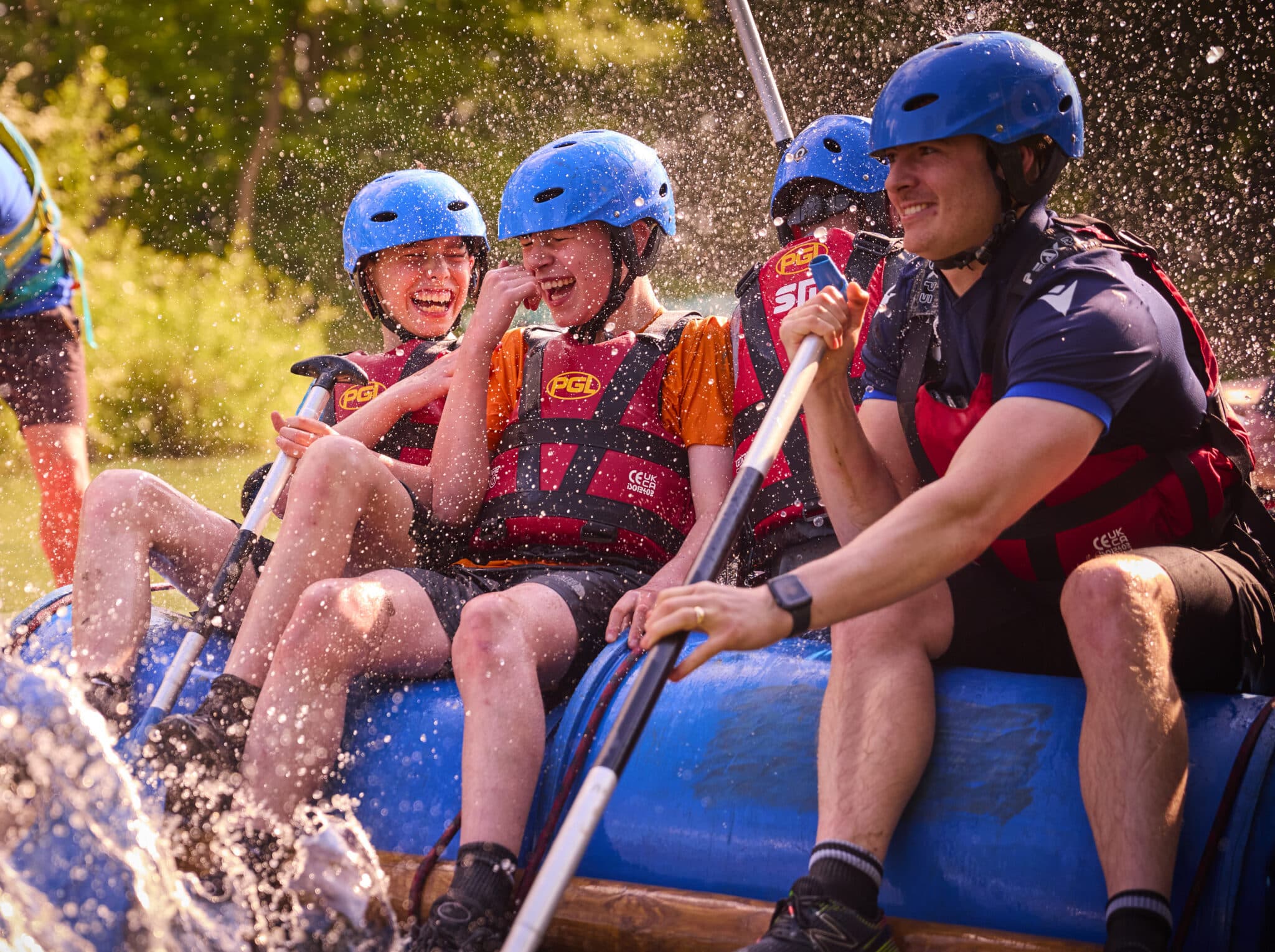 Four people wearing helmets and life jackets are white-water rafting, smiling and laughing as water splashes around them.