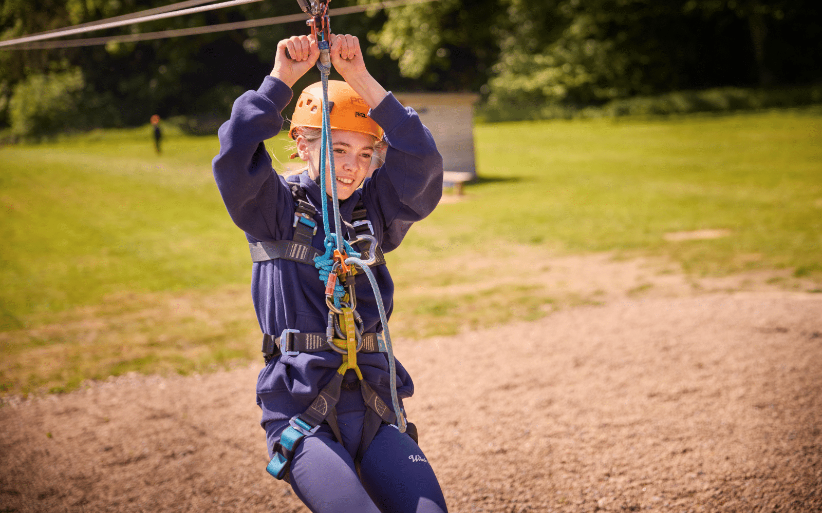 Person wearing a helmet and harness rides a zip wire over a gravel area in an outdoor setting with green grass and trees in the background.