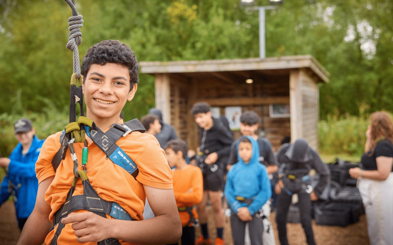 A smiling boy in an orange T-shirt and climbing harness stands at an outdoor ropes course, highlighting the benefits of outdoor learning.