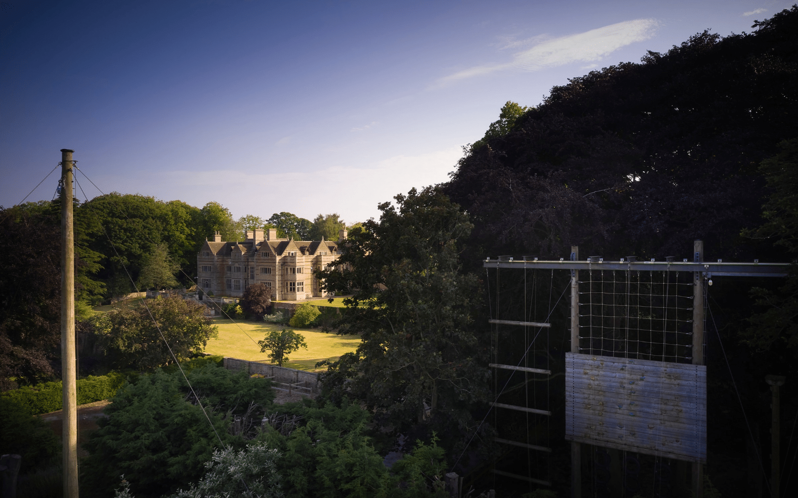 An historic manor house stands behind a large lawn, viewed from an elevated position next to an outdoor climbing wall at one of our school trip venues.