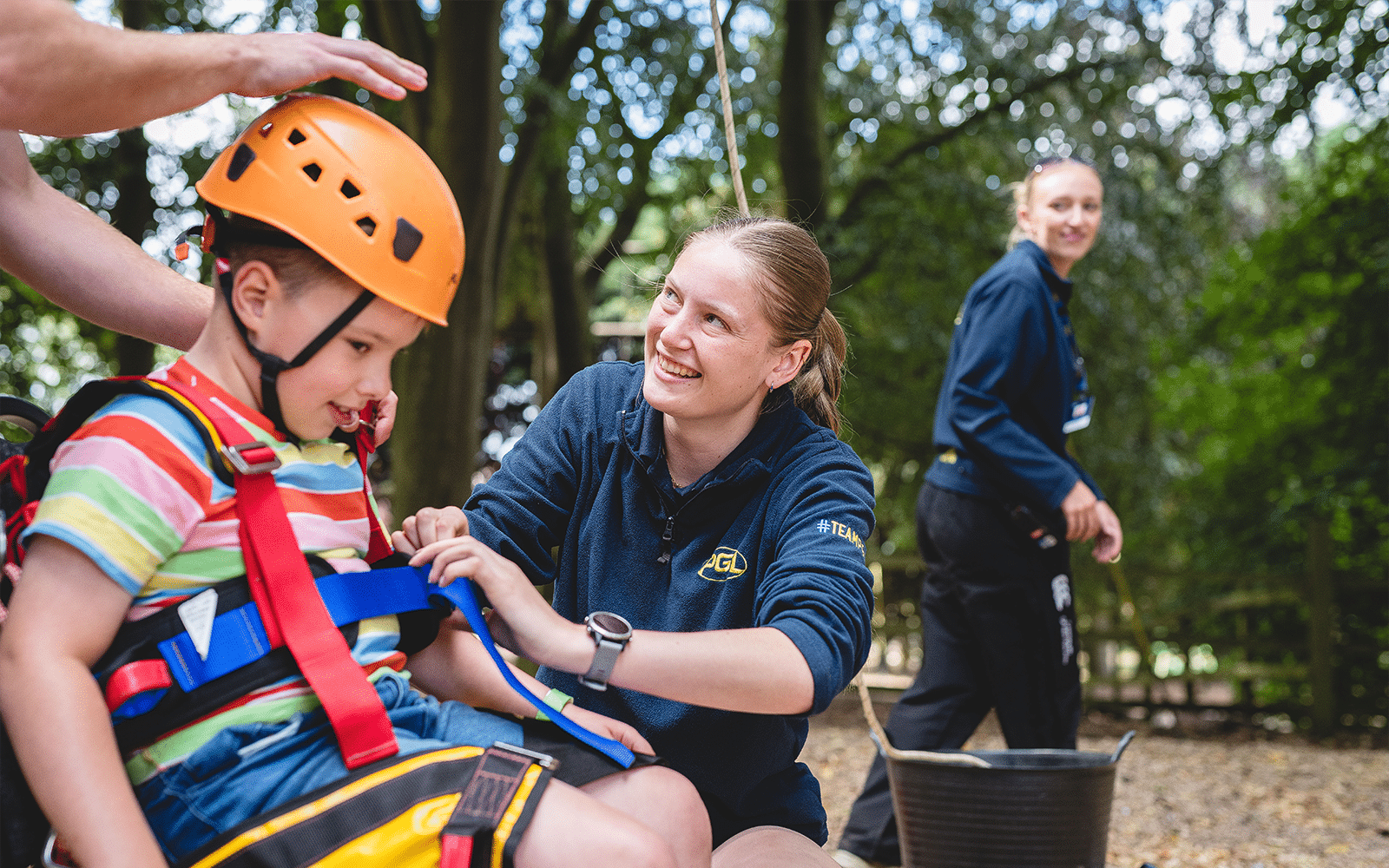 An instructor in a blue fleece smiles whilst helping a young boy in an orange climbing helmet and safety harness tighten his straps in a wooded area.