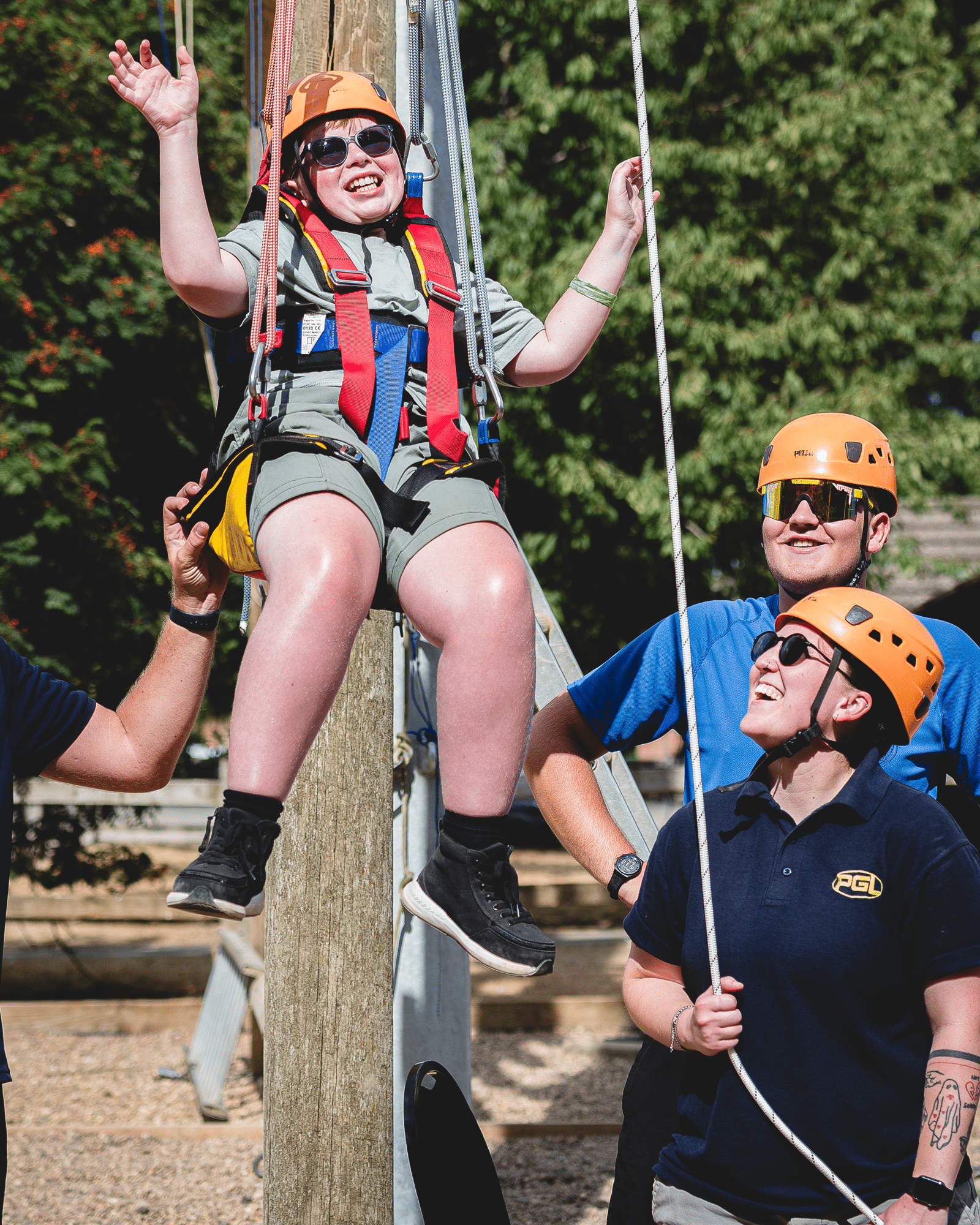 A person in a safety harness and helmet suspended from a high ropes course, assisted by two instructors on the ground.