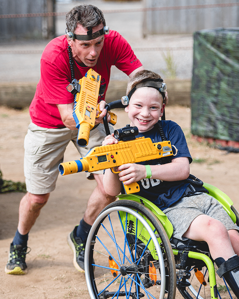 A man pushing a boy in a wheelchair, both wearing head-mounted sensors and holding yellow laser tag blasters.