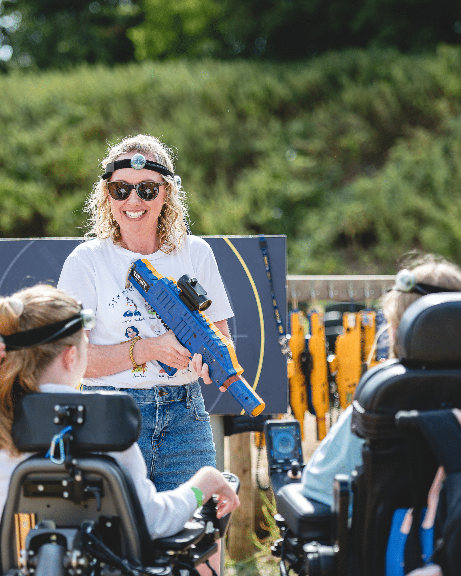 A woman smiling and holding a blue toy blaster stands between two people sitting in wheelchairs outdoors.