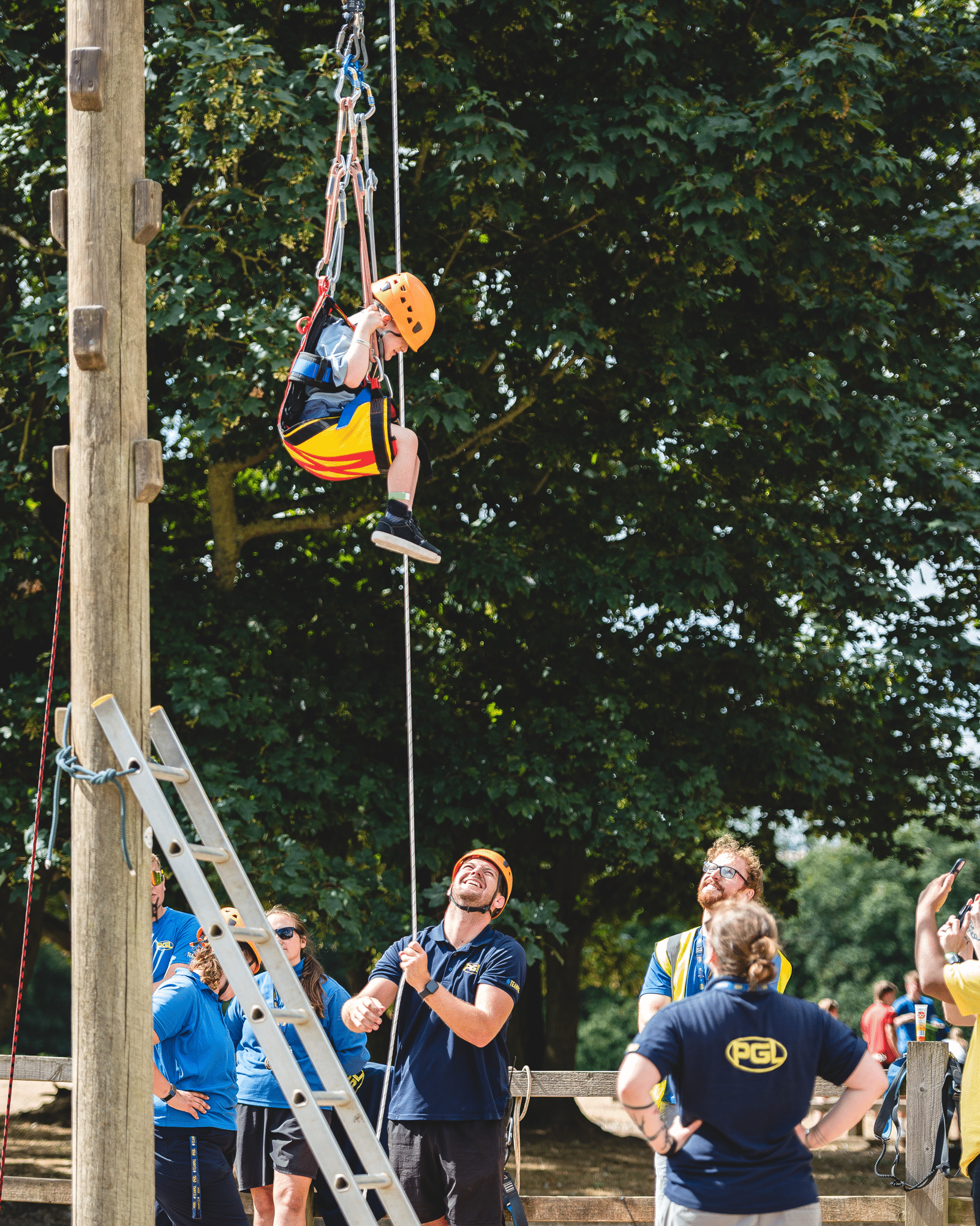 A child in a safety harness and helmet suspended by a rope from a high pole, with instructors watching from below.